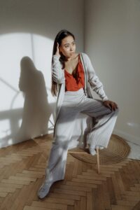 A woman in cozy loungewear sits thoughtfully on a stool in sunlight indoors.