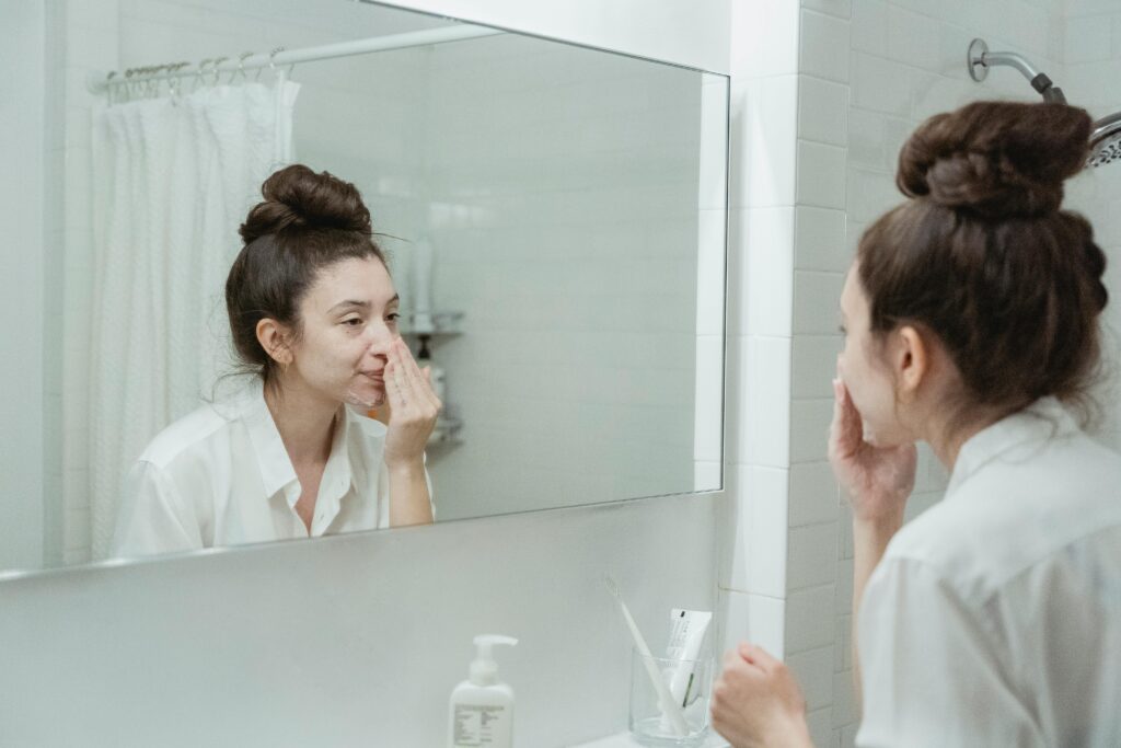 Caucasian woman in bathroom applying skincare product while looking in the mirror.